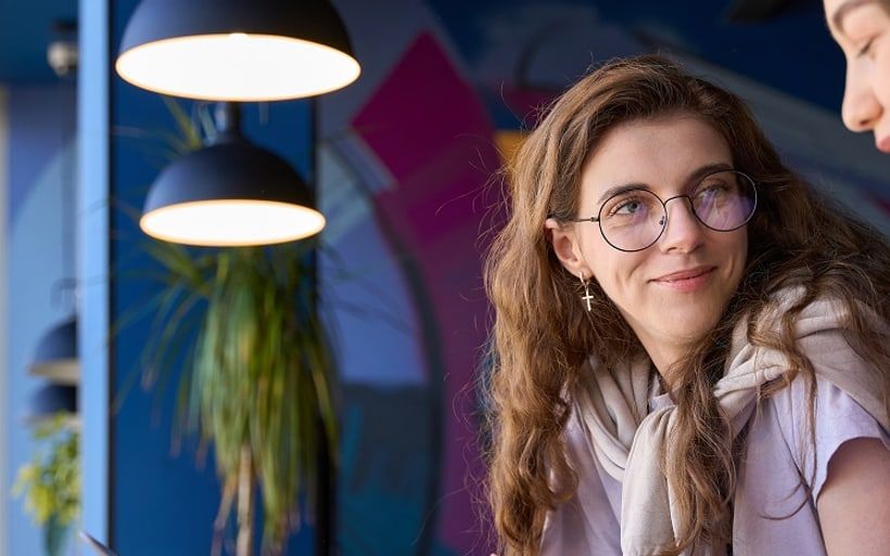 A woman in glasses turning to camera mid-conversation, pendant lamps behind her.