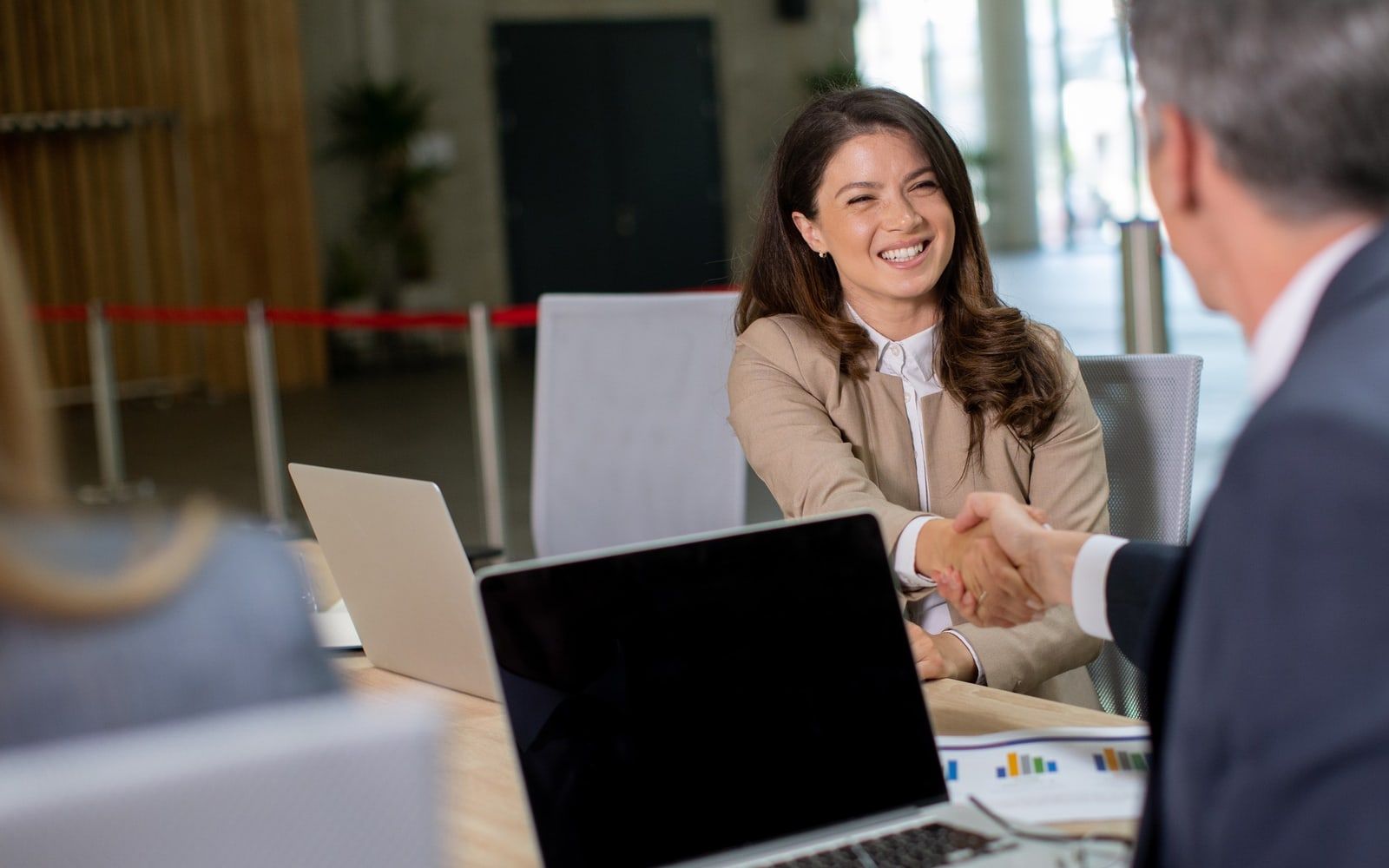 Two colleagues shaking hands across a meeting-room table, open laptops between them.