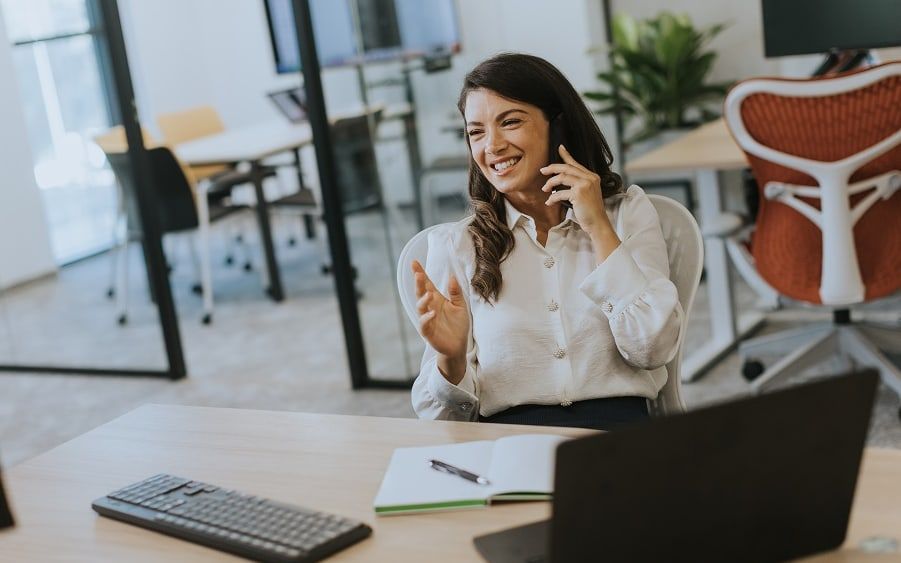 A woman on a call at her desk, gesturing as she talks, an open-plan office behind her.